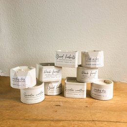 A collection of small, round, white scented candles in various fragrances including fresh linen, ocean, sandalwood, orange & cinnamon, lavender & chamomile, bergamot & spices, and more. The candles are presented in simple, minimalist packaging.
