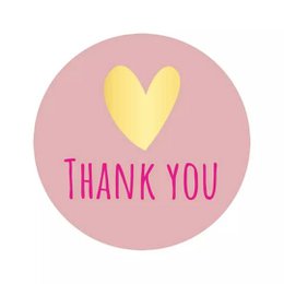 A round pink sticker with a yellow heart and the text "Thank You" in pink. The sticker appears to be adhered to a brown envelope or package.