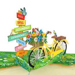 Colorful pop-up card with a bicycle, flowers, and directional signs depicting "Gesundheit", "Glück", and other positive messages.