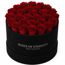 A large bouquet of vibrant red roses in a black box with the text "Roses of Eternity Amsterdam" printed on it. The roses appear to be high-quality, fresh, and arranged in a circular pattern.