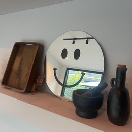A large round wall mirror with a smiling face design, made of black and white materials. The mirror is displayed on a shelf along with other decorative items.