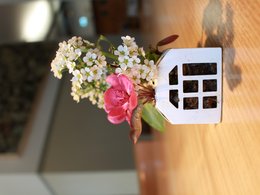 A white ceramic vase with a pink flower and small white flowers arranged inside. The vase has a simple, minimalist house-like shape.