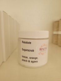 A white jar labeled "Resolute Sugar Scrub" with lemon, orange, and ginger scents.