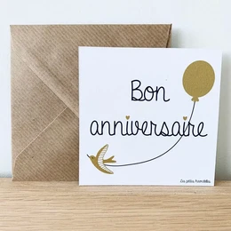 A white greeting card with the text "Bon anniversaire" (Happy birthday) and a gold balloon illustration. The card is accompanied by a brown envelope.