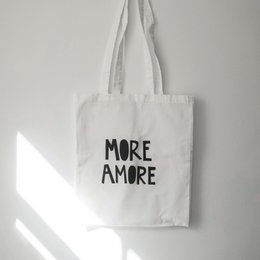 White canvas tote bag with the text "MORE AMORE" printed in black. The bag has a simple, minimalist design and appears to be a shoulder bag or reusable shopping bag.