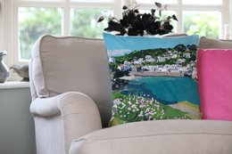 A plush, velvet cushion in a neutral beige color, featuring a scenic landscape of a coastal town with colorful flowers in the foreground.