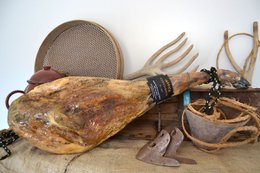 Large, cured Iberian ham with a dark brown exterior and visible marbling. Accompanied by traditional Spanish serving tools like a ham holder and carving knife.