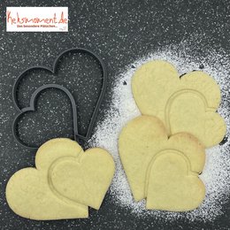 Two heart-shaped cookie cutters on a dark background with flour or sugar sprinkled around them. The cookie cutters are black in color and appear to be made of plastic or metal.
