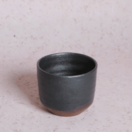 A small, cylindrical espresso cup made of dark-colored ceramic material. The cup has a simple, minimalist design with a smooth, matte finish.