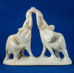 Soapstone statue depicting two kissing elephants. The elephants are carved in a stylized, abstract manner with smooth, rounded shapes. The statue has a neutral, off-white color.