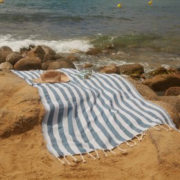 A striped beach towel or hammam towel in shades of blue and white, lying on the sand near the ocean with rocks in the background.
