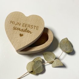 Wooden heart-shaped jewelry storage box with the text "Mijn eerste sieraadjes" (My first jewelry) engraved on it. The box contains small green stones or gems.