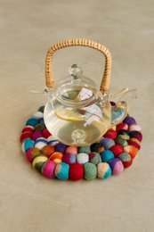Colorful round coaster with a glass teapot on top, surrounded by multicolored felt balls.