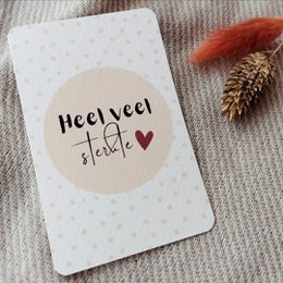 A small white greeting card with the text "Heel veel liefs" (Dutch for "Lots of love") and a heart symbol, set against a cozy, textured background with a pine cone.