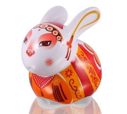 A porcelain piggy bank in the shape of a rabbit, featuring intricate red and gold designs. The rabbit has a round, plump body and large ears.