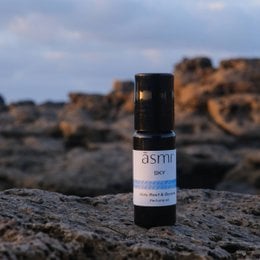 A small black glass bottle of essential oil labeled "ASMI" sits on a rocky surface. The bottle is accompanied by a white cylindrical container, also labeled "ASMI".