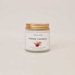 Transparent glass jar with gold lid containing a scented candle. The label features the text "Pomme cannelle" (cinnamon apple) and a small illustration of an apple.