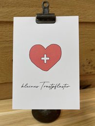 A small white postcard with a red heart-shaped symbol and the text "Bleiben Trostpflaster" (German for "Remain a comfort plaster") written below it. The postcard is clipped to a wooden surface with a black metal clip.