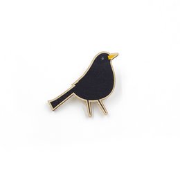 Wooden pin in the shape of a blackbird, a small dark-colored songbird. The pin features a simple, minimalist design with a black body and gold-colored accents.