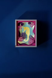 Colorful framed artwork depicting a whimsical mermaid figure against a tropical background with palm trees. The mermaid has vibrant green skin and is surrounded by pink and purple accents, creating a playful and imaginative scene.