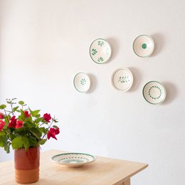 Decorative wall plates in various designs, including circular patterns and floral motifs. The plates are displayed on a wooden shelf, complemented by a vase of red roses.

The image shows a set of decorative wall plates in various designs. The plates feature circular patterns and floral motifs, with colors ranging from white to green. The plates are displayed on a wooden shelf, creating a visually appealing arrangement. A vase of red roses is placed in front of the plates, adding a touch of natural beauty to the scene.