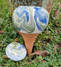 A round, tall ceramic planter with a blue and green wave pattern. The planter is sitting in a cardboard box on the ground surrounded by grass and leaves.