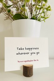 A greeting card with the text "Take happiness wherever you go. Never apologize for it." The card features a simple, minimalist design with the text centered on a light-colored background.