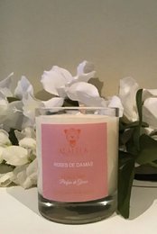 Scented candle with rose de damas fragrance, presented in a pink glass container with a decorative label. The candle has a burn time of 25 or 55 hours depending on the size variant.