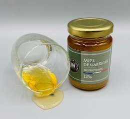 A glass jar of golden honey labeled "Miel de Garrigue 125g : L'Essence de la Provence en Pot" with a glass dipper beside it. The honey appears to be a natural, artisanal product from the Provence region of France.