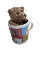 A plush teddy bear sitting in a colorful mug with a cartoon design. The mug appears to be made of ceramic or porcelain.