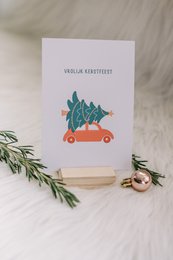 A Christmas card featuring a red car carrying a Christmas tree on its roof. The card has a minimalist, illustrated design with the text "Merry Christmas" in a simple, elegant font.