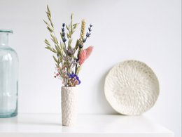 A tall, cylindrical vase containing a bouquet of colorful dried flowers and foliage, including pink and purple blooms. The vase has a textured, off-white ceramic surface.