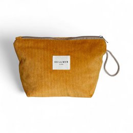 A large, rectangular makeup pouch made of ribbed, mustard-colored corduroy material. The pouch has a zipper closure and a small fabric tag with the brand name "Baggu" sewn onto it.