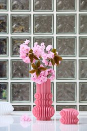  A tall, pink ceramic vase holding a bouquet of pink flowers, placed on a table with pink chairs in front of a glass window.