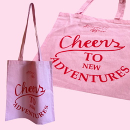 A pink cotton tote bag with the text "Cheers to New Adventures" printed in red. The bag appears to be made of a durable, natural fabric like jute or canvas.