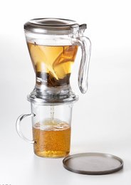 A clear glass teapot with a metal lid, filled with a golden-colored tea. The teapot has a handle and a spout, and is sitting on a clear glass surface.