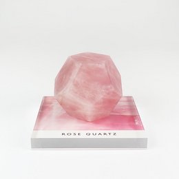 A large, faceted rose quartz crystal on a white display stand. The crystal has a soft, pink hue and a geometric, polyhedral shape.