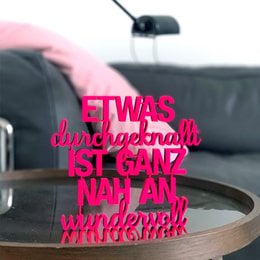 A decorative pink sign with the text "Es war einmal ist ganz nah an wunderbar" (Once upon a time is very close to wonderful). The sign appears to be made of plastic or a similar material and is displayed on a surface with a plant in the background.