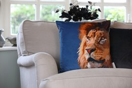 A large, fluffy lion-themed cushion with a close-up image of a lion's face on a blue background, sitting on a beige couch in a cozy living room setting.