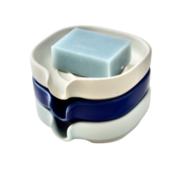 A self-draining porcelain soap dish in matte white, glossy cobalt blue, and glossy celadon blue colors. The dish has a unique shape with a raised center platform to allow water to drain away from the soap.