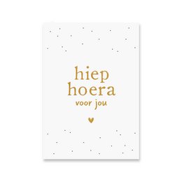 A white greeting card with gold foil text that reads "Hiep hoera voor jou" (Hooray for you in Dutch). The card has a simple, minimalist design with small gold dots scattered across the background.