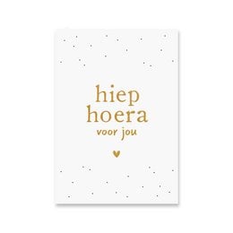 A white greeting card with gold foil text that reads "Hiep hoera voor jou" (Hooray for you in Dutch). The card has a simple, minimalist design with small gold dots scattered across the background.