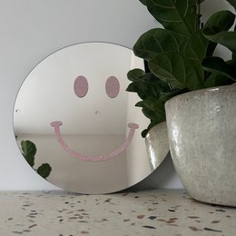 A round wall mirror with a large pink smiley face design on a black background. The mirror is placed on a table next to a potted plant.