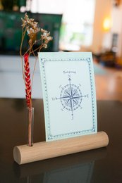 A small glass vase with a card holder feature, featuring a compass design on the card. The vase has a wooden stand and contains dried floral elements.