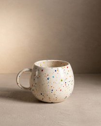 A round, white ceramic mug with a speckled pattern of colorful dots, suitable for parties and casual use.