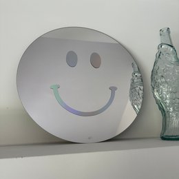 A round, silver-colored wall mirror with a holographic smiley face design. The mirror has a simple, minimalist style and appears to be made of a reflective material.