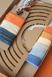 Colorful DIY rainbow craft kit with vintage-inspired design. Includes wooden rainbow shape, yarn in various shades of blue, orange, and white, and instructions.
