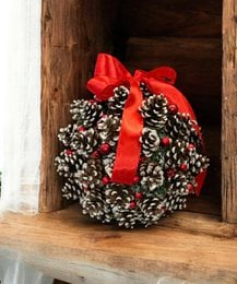 A round, handmade Christmas ornament made of pine cones, with a red satin bow. The ornament is approximately 20-23 cm in diameter and comes in various color variants including pink, multicolored, gold, and red.