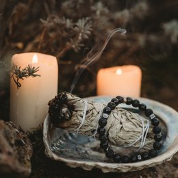 This intention bracelet features a silver charm with a leaf design, black beads, and a white candle with a leaf motif. The bracelet has a natural, earthy style that would complement a variety of outfits.
