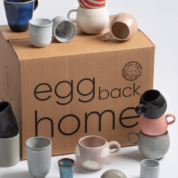 A set of 24 assorted mugs in various colors, shapes, and designs, including solid colors, patterns, and text. The mugs are packaged in a cardboard box labeled "egg back home".